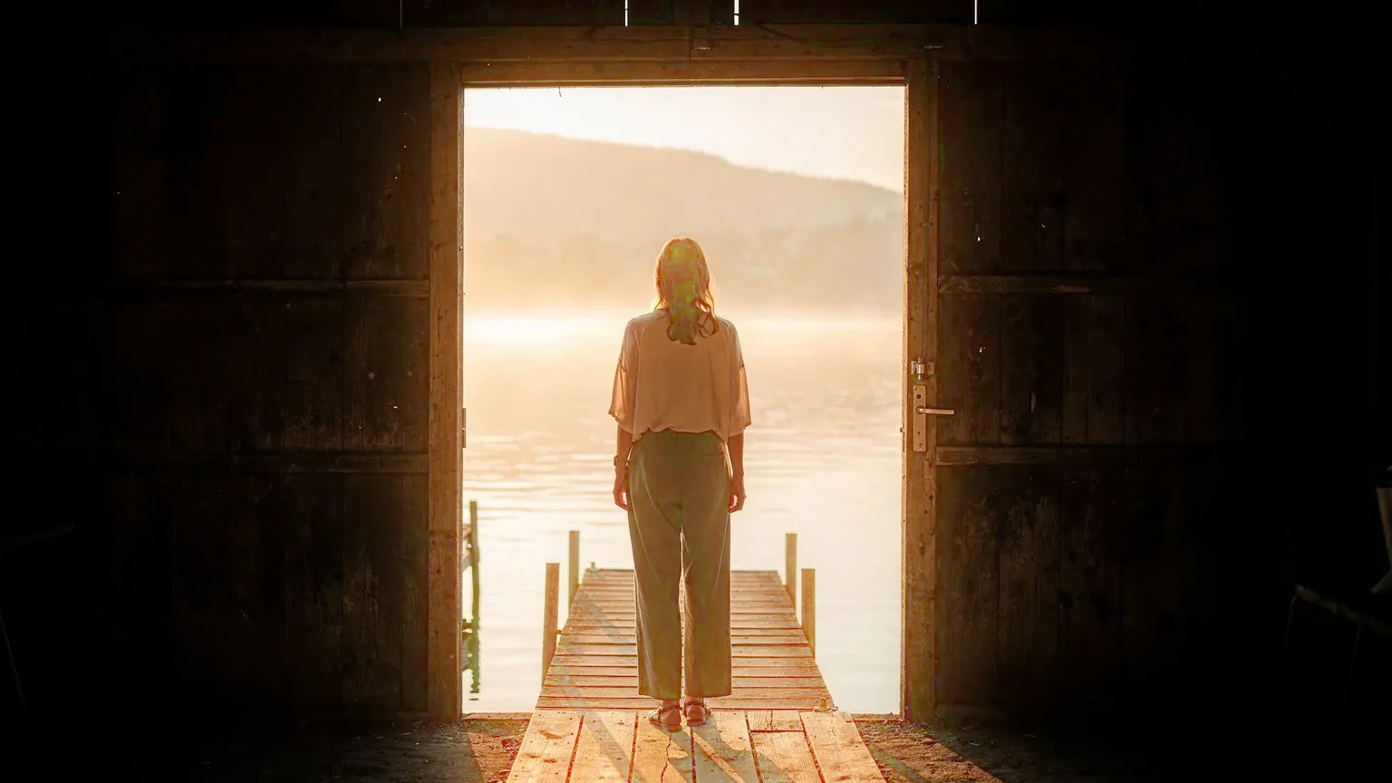 Selbstbewusstsein Coaching am Bodensee: Frau steht aufrecht in der offenen Tür eines Bootshauses, davor der Steg und der See im Morgenlicht (KI-generiertes Bild)